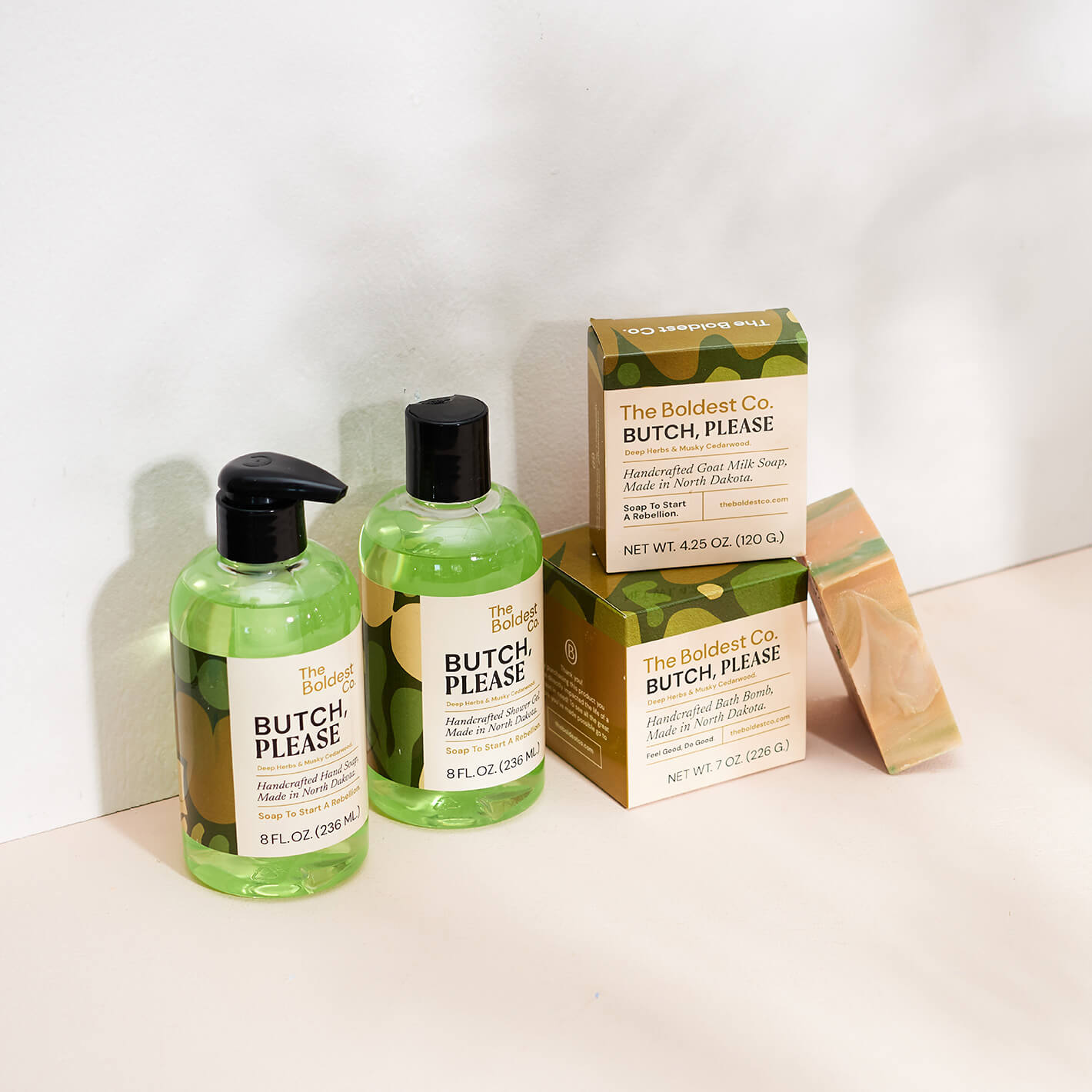 Brand identity case study image of The Boldest co’s ‘Butch, Please’ scent range. A hand wash, shower gel, soap box and bath bomb box feature green detailing and sit in a cream coloured background.