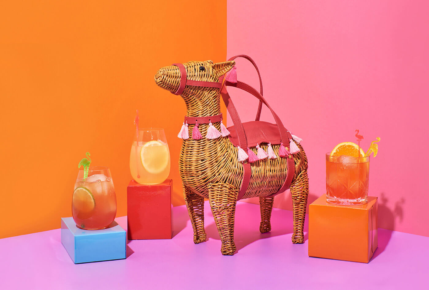 Bright orange and pink scene with llama shaped wicker handbag surrounded by cocktails