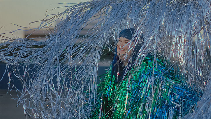 A blue haired woman in a green tinsel jacket laughs amidst a silver tinsel wall on a roof top as the sun sets in the background.