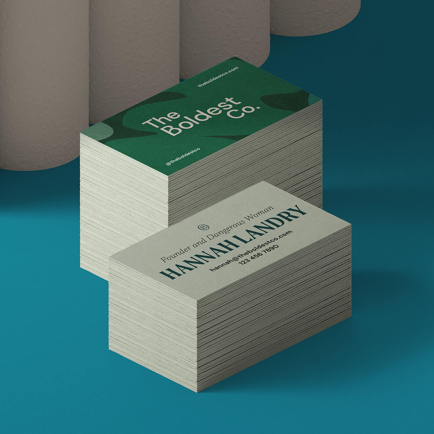 The Boldest Co business cards sit in a deep green background. The front of the card shows the brand logo on a green blob-textured background while the back features founder Hannah’s details and simple typesetting.