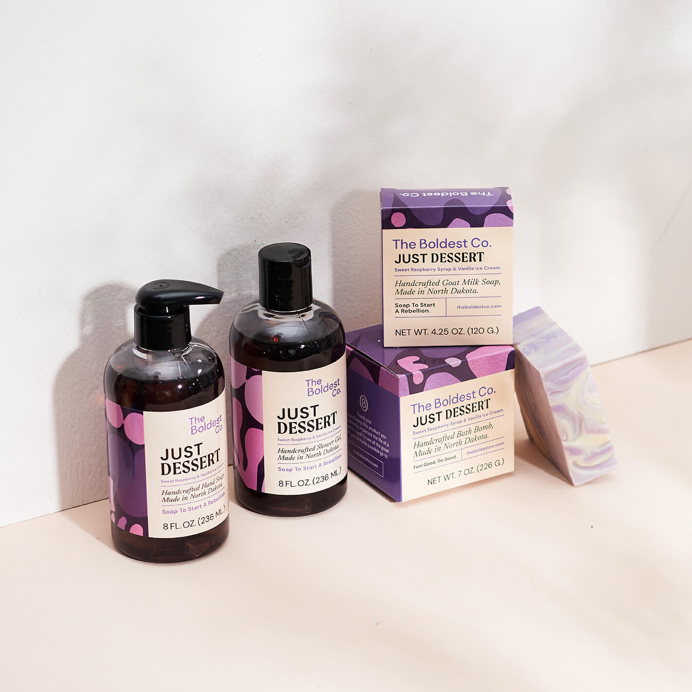 Brand identity case study image of The Boldest co’s ‘Just Dessert’ scent range. A hand wash, shower gel, soap box and bath bomb box feature purple detailing and sit in a cream coloured background.