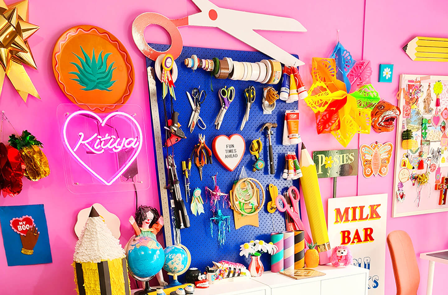 Brand identity case study image showing Kitiya Palaskas’ colourful studio space. From neon and vintage signs, paper ornaments, a blue pegboard and the bright pink walls, the space feels lively.
